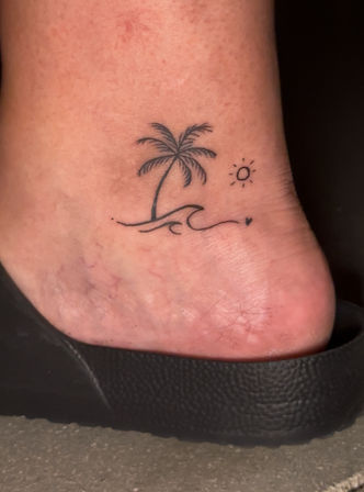 Ankle tattoo of a minimalist palm tree and wave with a small sun and tiny heart, shown on skin above a black slide sandal