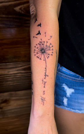 Close-up of a forearm with a fine-line black dandelion tattoo; seeds and bird silhouettes blowing upward, stem flowing into cursive script reading "until we meet again".