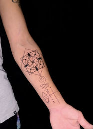Inner forearm black-ink geometric mandala tattoo with compass-arrow accents, spiral and abstract line-and-triangle symbols on light skin, close-up against black background