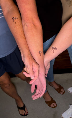 Three linked forearms and hands displaying matching small minimalist black bow outline tattoos on inner forearms, casual summer clothing and sandals