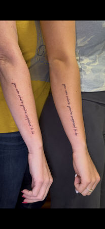 Close-up of two people’s inner forearms side-by-side with matching black cursive forearm tattoos reading 'you are where you're supposed to be', vertical script and casual clothing in the background.