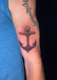 Close-up of a person's inner forearm with a freshly inked black-shaded nautical anchor tattoo, faint redness around the design, barbed-wire tattoo above and a blue shirt sleeve at the edge.