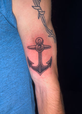 Close-up of a person's inner forearm with a freshly inked black-shaded nautical anchor tattoo, faint redness around the design, barbed-wire tattoo above and a blue shirt sleeve at the edge.