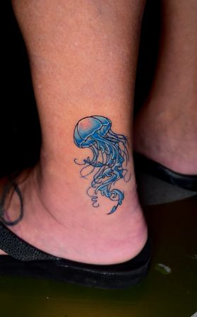 Tiny blue jellyfish tattoo with flowing curly tentacles on the inner ankle above a black sandal.