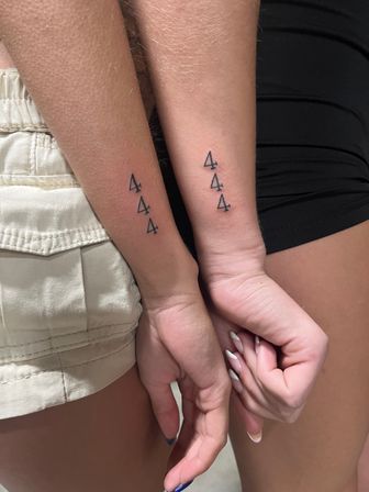 Close-up of two people with matching minimalist tattoos: three small black outlined number 4s stacked vertically on each inner forearm, hands held close together, casual shorts and black skirt visible.