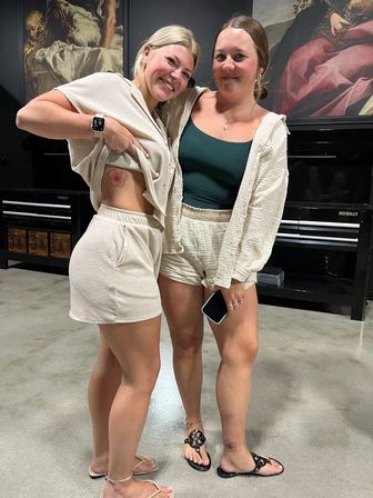 Two smiling friends in beige loungewear posing indoors; one lifts her top to show a red flower rib tattoo while the other shows a small ankle flower tattoo, both in sandals in front of framed artwork.