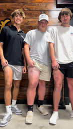 Three young men in shorts and sneakers showing matching small sun-and-wave thigh tattoos inside a wood-paneled tattoo studio.