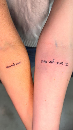 Close-up of two inner forearms with matching minimalist black-ink script tattoos reading "I love you more," simple cursive matching tattoos on tanned and fair skin.