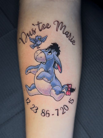Colorful forearm tattoo of Eeyore (Winnie the Pooh) with a small bluebird above and pink tail bow, framed by cursive name script and a semi-circle of black numbers.