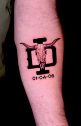 Forearm tattoo of a longhorn skull centered on a bold black emblem, western vibe, with the date 01-04-06 beneath.