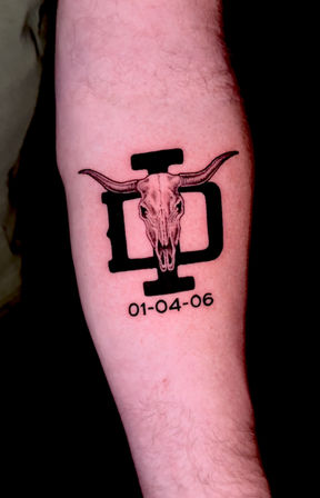 Forearm tattoo of a longhorn skull centered on a bold black emblem, western vibe, with the date 01-04-06 beneath.