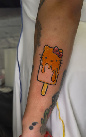 Playful inner-forearm tattoo of a melting cartoon kitty popsicle with a red bow, bright orange and yellow ink