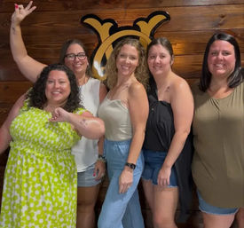 Five smiling women in casual summer outfits posing for a group photo in front of a wood-paneled wall with a decorative sign, showing small shoulder and forearm tattoos — friends' night out at a casual bar or restaurant.