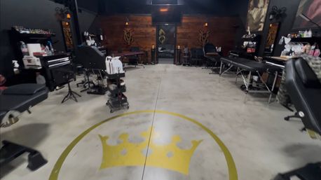 Spacious tattoo studio interior with concrete floor and large gold crown logo, black tattoo chairs and treatment tables along the sides, rolling tool carts, and a warm wood accent wall.