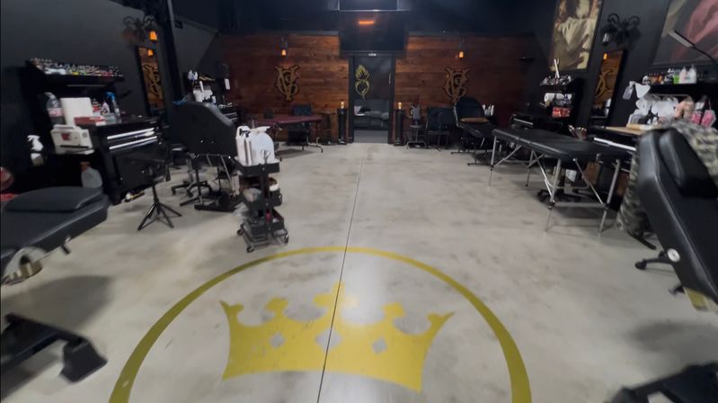 Spacious tattoo studio interior with concrete floor and large gold crown logo, black tattoo chairs and treatment tables along the sides, rolling tool carts, and a warm wood accent wall.