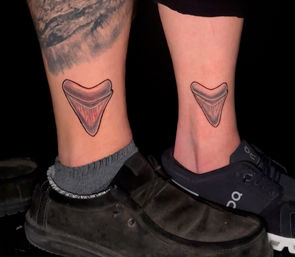 Close-up of two ankles with matching stylized shark-tooth ankle tattoos — black outline with pink-gray shading and vertical texture lines, casual black shoes and dark background.