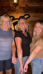 Three smiling women in casual clothes pose against a wood‑paneled wall with a hanging lantern and stylized sign, showing matching tiny bow tattoos on their forearms — friendly group photo.