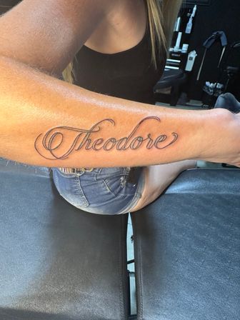 Close-up of a fresh cursive forearm tattoo reading "Theodore" in elegant black script on a person's inner forearm, photographed in a tattoo studio setting.