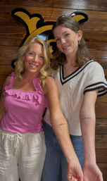 Two smiling women at a tattoo studio pose against a wooden wall with a black-and-yellow sign, showing matching small script tattoos on their inner forearms while wearing casual summer tops.