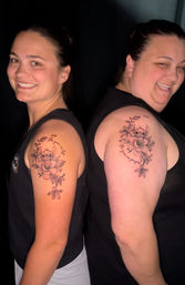 Two smiling women standing back-to-back showing matching upper-arm floral tattoos of Stitch peeking from lilies and leaves with cursive script, dark studio background