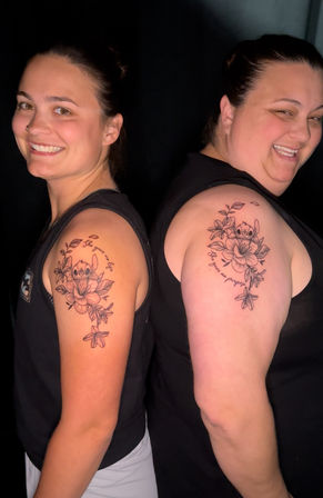 Two smiling women standing back-to-back showing matching upper-arm floral tattoos of Stitch peeking from lilies and leaves with cursive script, dark studio background