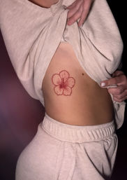 Close-up of a person lifting a beige sweater to reveal a red hibiscus flower tattoo on the side rib area, white-painted nails and matching beige lounge pants visible.