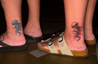 Close-up of two ankles with matching jellyfish tattoos—one blue, one black ink—above sandals and flip-flops on a dark floor