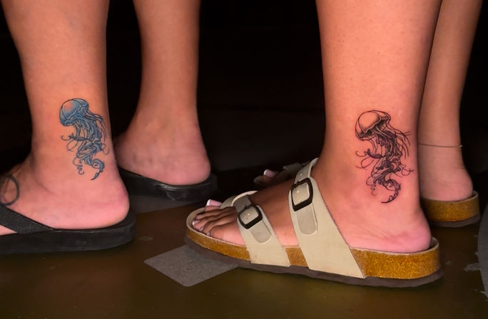 Close-up of two ankles with matching jellyfish tattoos—one blue, one black ink—above sandals and flip-flops on a dark floor
