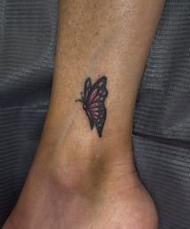 Close-up of a small pink and black butterfly tattoo on an ankle with a grey textured fabric background