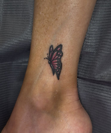 Close-up of a small pink and black butterfly tattoo on an ankle with a grey textured fabric background