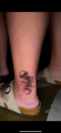 Close-up of a delicate black linework jellyfish tattoo on the inner ankle above a beige sandal.