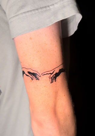 Close-up of a black-ink armband forearm tattoo showing two stylized hands reaching toward each other in minimalist, Michelangelo-inspired linework.