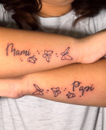 Matching black-ink forearm tattoos reading “Mami” and “Papi” in cursive, linked by minimalist line-art butterflies and tiny star dots — delicate family tribute near the wrists.