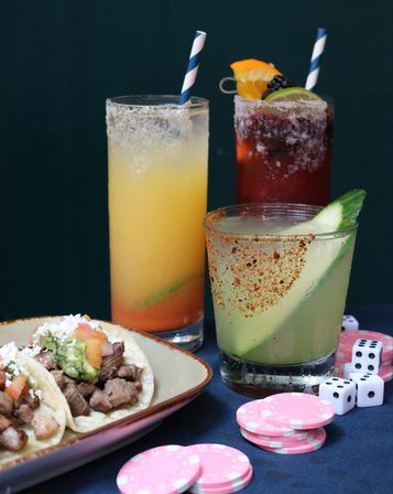 Mexican-inspired spread with two tall citrus cocktails and a cucumber-chili rimmed margarita, carne asada tacos topped with guacamole and queso fresco, and pink poker chips and dice on the table.
