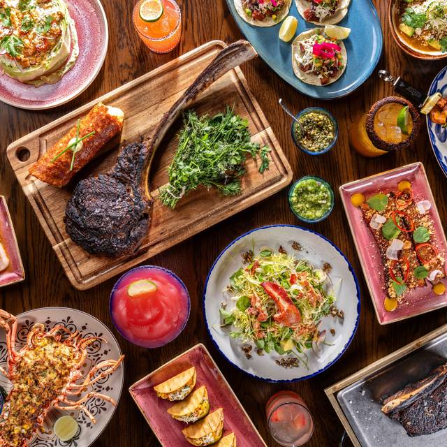 Vibrant overhead restaurant spread on a wooden table: charred tomahawk steak on a cutting board, lobster, mini tacos, salads, colorful appetizers, dips and bright cocktails — a festive seafood and steak feast