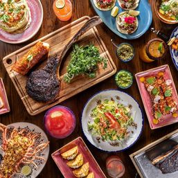 Vibrant overhead restaurant spread on a wooden table: charred tomahawk steak on a cutting board, lobster, mini tacos, salads, colorful appetizers, dips and bright cocktails — a festive seafood and steak feast