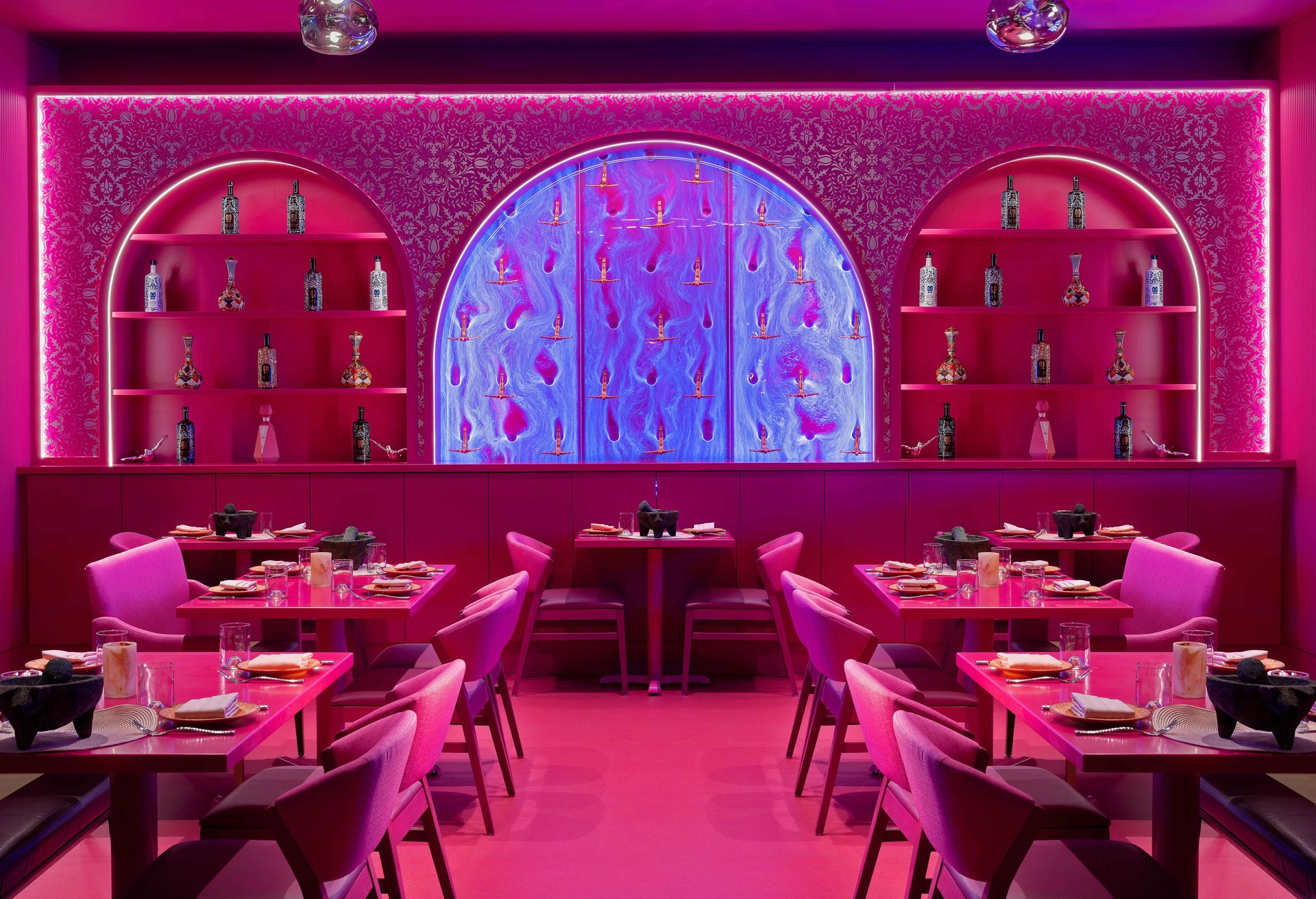Vibrant pink restaurant interior with neon-lit arched shelves displaying bottles, a glowing blue central art panel, and neatly set tables and chairs