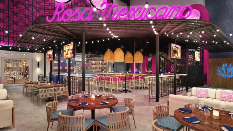 Vibrant modern Mexican restaurant interior with pink neon script sign above a covered pergola, string lights and wicker pendant lamps, central bar with high stools, round wooden dining tables set with blue napkins and comfortable lounge seating.