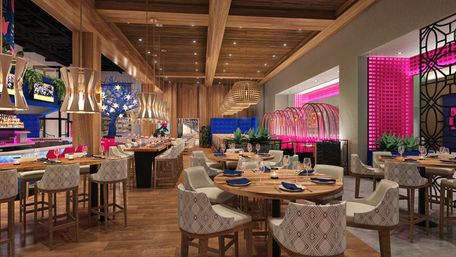 Upscale modern restaurant interior with wooden beams and floor, pendant-lit bar, round dining tables with patterned chairs, blue banquette seating and vibrant pink neon-lit lounge area.
