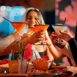 Lively group toasting colorful margarita-style cocktails over a flower-decorated restaurant table with appetizers and festive lights