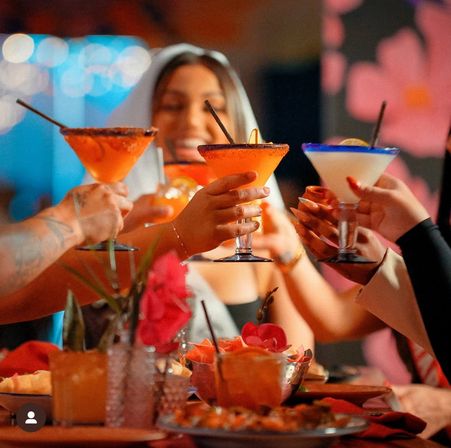 Lively group toasting colorful margarita-style cocktails over a flower-decorated restaurant table with appetizers and festive lights