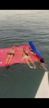 Three people relaxing on a large red floating mat in calm open sea while a fourth person holds on from the water beside a rolled blue mat and a white boat bow visible in the corner.
