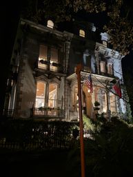 Night view of a grand Victorian mansion facade aglow with warm interior lights, ornate iron balconies, columned entrance and an American flag by the doorway.