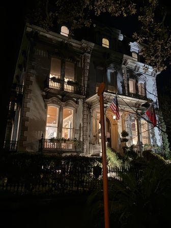 Night view of a grand Victorian mansion facade aglow with warm interior lights, ornate iron balconies, columned entrance and an American flag by the doorway.