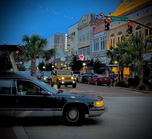 Evening downtown scene on Drayton St — palm trees, string lights, historic storefronts, traffic with a yellow Jeep and a black car carrying a skeleton prop