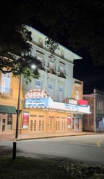 Night view of a historic downtown theater with a glowing vintage marquee casting warm light on an ornate three‑story façade and closed doors along a tree‑lined main street.