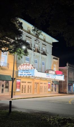 Night view of a historic downtown theater with a glowing vintage marquee casting warm light on an ornate three‑story façade and closed doors along a tree‑lined main street.
