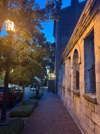 Dusk in a historic downtown: brick sidewalk beside a stone arched facade lit by a warm streetlamp, trees lining the street, parked cars and an American flag under a deep blue sky.