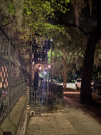 Nighttime southern streetscape with ornate wrought-iron balcony and brick facade, warm street lamps, Spanish moss-draped oak trees and a silhouetted figure on the sidewalk near parked cars.