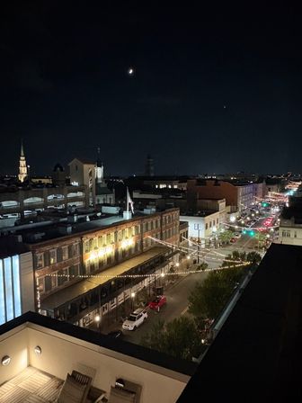 Rooftop view of a historic downtown main street at night with brick storefronts and twinkling string lights over the road, parked cars below, an illuminated church spire on the skyline, and a crescent moon overhead.